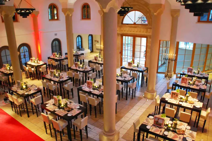 Das Bild zeigt einen elegant dekorierten Speisesaal mit mehreren Tischen, die für ein Abendessen vorbereitet sind. Die Tische sind mit weißen Tischdecken, Geschirr, Gläsern und kleinen Dekorationen ausgestattet, was auf ein bevorstehendes Event wie ein Bankett oder eine Gala hinweist. Die Beleuchtung ist warm und einladend, und die roten Akzente, wie der rote Teppich und die beleuchteten Säulen, geben dem Raum eine festliche Atmosphäre. Es ist ein repräsentativer Raum, der für feierliche Anlässe geeignet scheint.