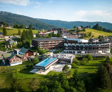 Panorama eines Resorts in alpiner Landschaft, umgeben von grünen Wiesen und Wäldern. Das moderne Hotel verfügt über einen beeindruckenden Infinity-Pool und großzügige Terrassenbereiche, eingebettet in eine charmante Bergdorf-Kulisse.