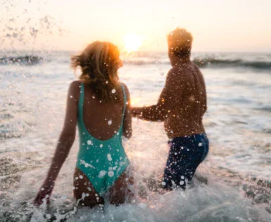 Eine Frau in türkisem Badeanzug und ein Mann in blauer Badehose planschen bei Sonnenuntergang im Meer. 