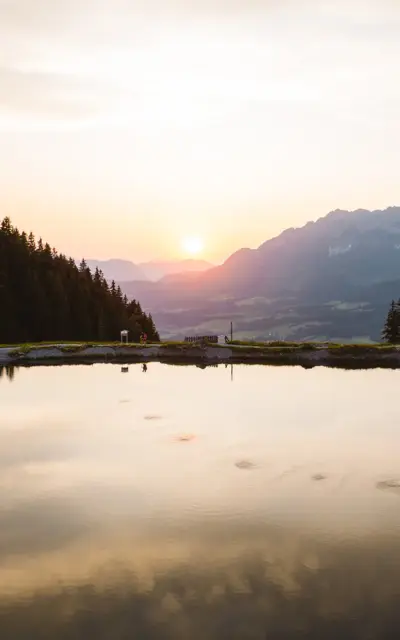 Ein ruhiger See mit Bäumen und Bergen im Hintergrund, reflektiert im Wasser.