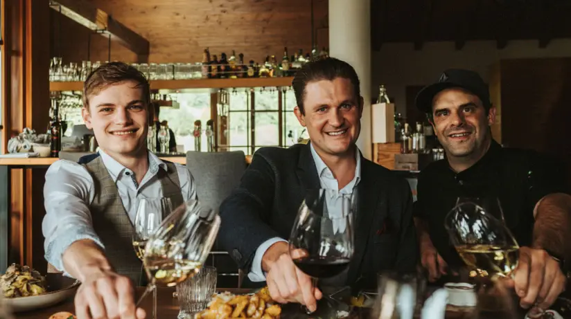 Drei Männer sitzen an einem Tisch in einem Restaurant und stoßen mit Weingläsern an. Sie lächeln in die Kamera und wirken fröhlich. Im Hintergrund ist eine Bar mit Flaschen und Gläsern zu sehen. Auf dem Tisch stehen verschiedene Gerichte.