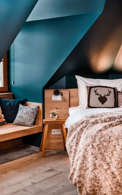 Modern gestaltetes Schlafzimmer mit geometrischen blauen Wandelementen und Holzmöbeln. Ein gemütliches Bett mit hochwertiger Bettwäsche und dekorativen Kissen lädt zum Entspannen ein. Natürliches Licht fällt durch das Fenster ein und erhellt den Raum, während die Kombination aus warmen Holztönen und kühlen Blautönen eine ruhige und einladende Atmosphäre schafft.