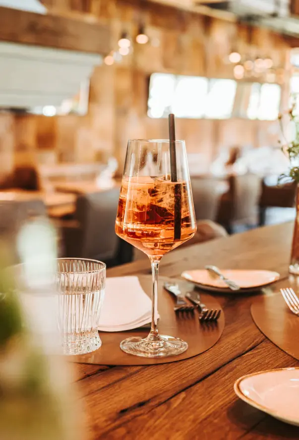 Ein Glas Roséwein mit einem Strohhalm, präsentiert auf einem Holztisch in einem stilvollen Restaurant. Der Fokus liegt auf dem Getränk im Vordergrund, während das warme Ambiente des Lokals mit unscharfen Sitzgelegenheiten und sanfter Beleuchtung im Hintergrund eine gemütliche und einladende Atmosphäre schafft."