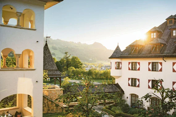  Die Morgensonne scheint hinter dem Hotel in Kitzbühel mit traditioneller Architektur hervor, das in eine idyllische, hügelige Landschaft eingebettet ist
