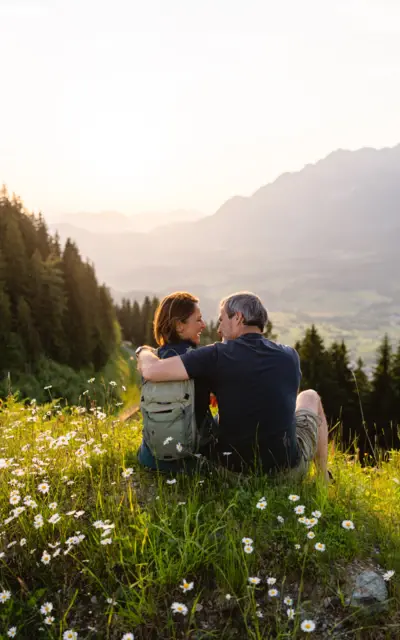 Ein Paar sitzt auf einer blühenden Bergwiese und schaut in die Ferne. Sie umarmen sich und genießen die Sommersonne über den malerischen Bergen und dem Tal im Hintergrund.