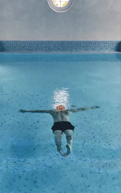 A man swims in the blue indoor pool with his arms outstretched. 