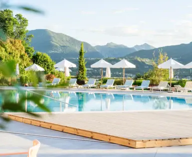 Outdoor pool surrounded by sun loungers and parasols, flanked by trees under a blue sky.