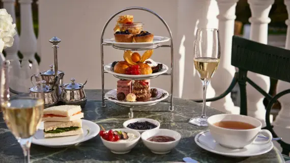 Ein stilvoll gedeckter Tisch für Afternoon Tea auf einer Terrasse mit weißem Geländer. Eine dreistöckige Etagere präsentiert Gebäck, Croissants und Pralinen.