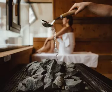 Ein Aufguss wird in einer Sauna gemacht, wobei Wasser aus einer Holzkelle über heiße Steine gegossen wird, was Dampf erzeugt. Im Hintergrund sitzt entspannt eine Person, umhüllt von einem Handtuch, in einer hölzernen Saunalandschaft.