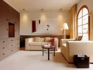 A bright living area with two beige sofas. There is a large wooden wall unit on the left and high, rounded window fronts on the right.