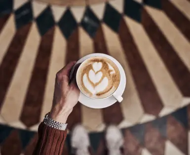 Eine Hand hält eine Tasse Cappuccino mit kunstvoller Latte Art in Herzform. Im Hintergrund ein geometrisch gemusterter Boden in warmen Braun- und Beigetönen. 