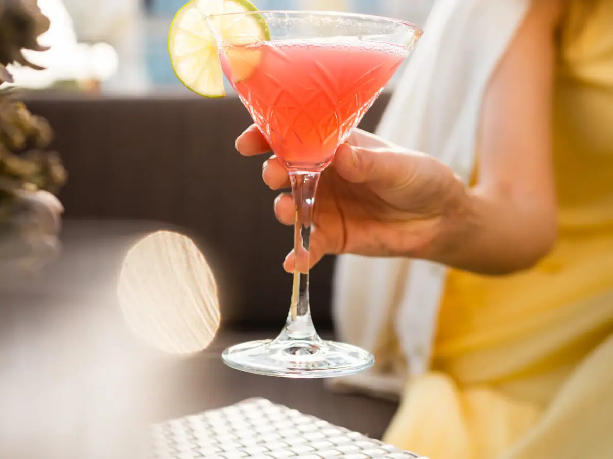 Eine Frau hält ein Glas mit einem rosa Cocktail auf einer Terrasse.