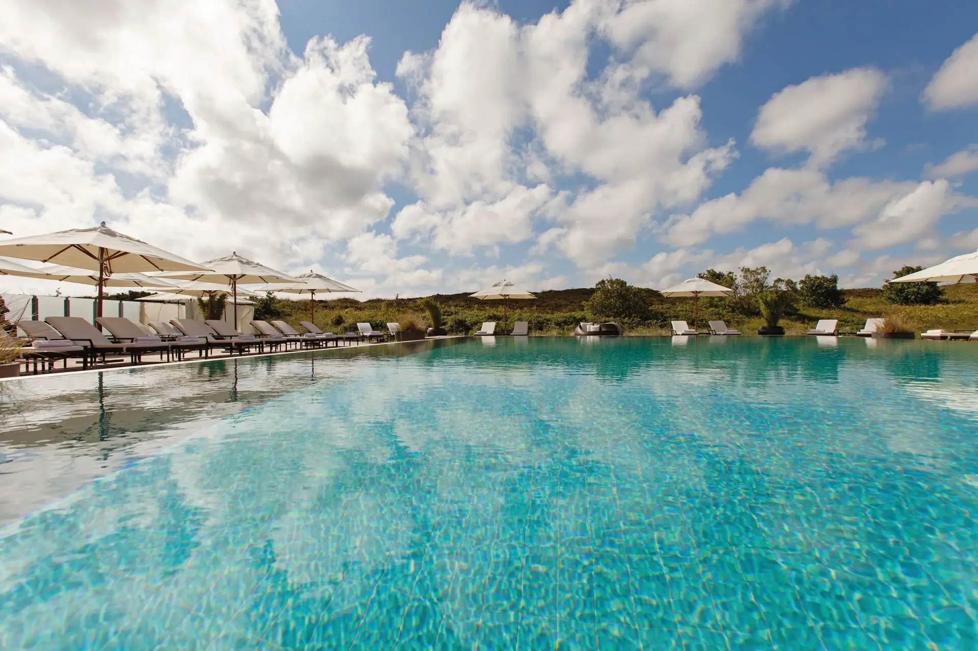 Außenpool A-ROSA Sylt Ein Pool mit Liegestühlen und Sonnenschirmen und im Hintergrund sind Dünen zu sehen.