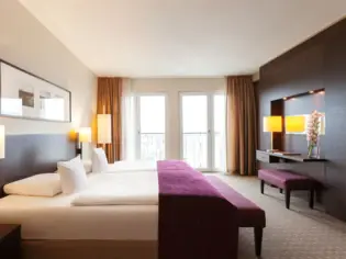 A hotel room with a large bed on which a purple bedspread lies. Opposite is a large window front with orange curtains. The wall on the left is covered with dark wooden panels.