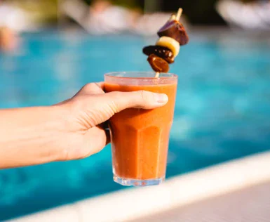 Eine Hand hält ein Glas mit einem erfrischenden, orangefarbenen Smoothie, garniert mit einer Spieß aus Obst und Schokolade, vor dem Hintergrund eines lebhaften Außenpools.
