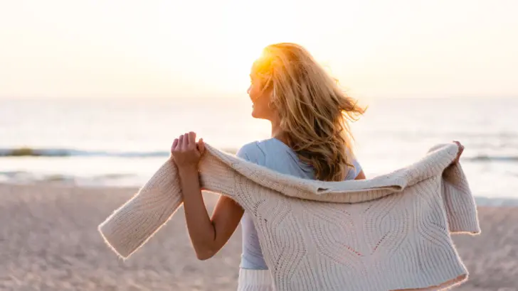 Frau hält einen Pullover bei Sonnenuntergang am Strand.