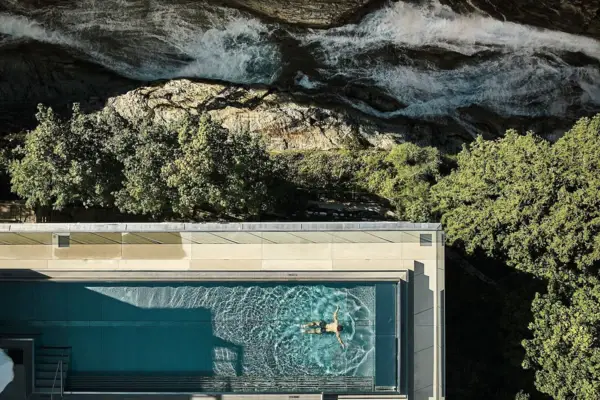 Es wurde ein länglicher Outdoor-Pool von oben aus der Vogelperspektive fotografiert, in dem ein Mann schwimmt. Zudem ist im oberen Teil des Bildes ein Wasserfall zu sehen.