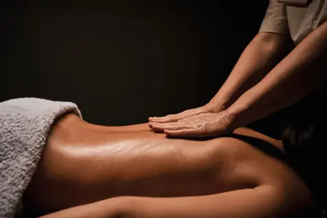 Eine Frau liegt auf einer Massageliege und ist mit einem Handtuch bedeckt. Auf ihrem Rücken ruhen zwei Hände, die sie massieren.