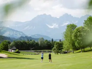 Zwei Personen auf einem gepflegten Golfplatz mit Blick auf eine beeindruckende Alpenkulisse, umgeben von grünen Bäumen und einer idyllischen Landschaft unter einem teilweise bewölkten Himmel.