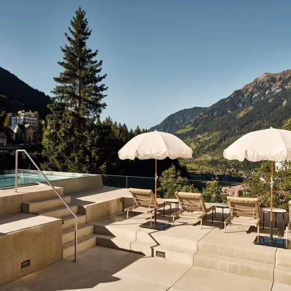 Sonniger Poolbereich mit Sonnenschirmen und Liegestühlen, umgeben von Bäumen und Gebirge im Hintergrund.