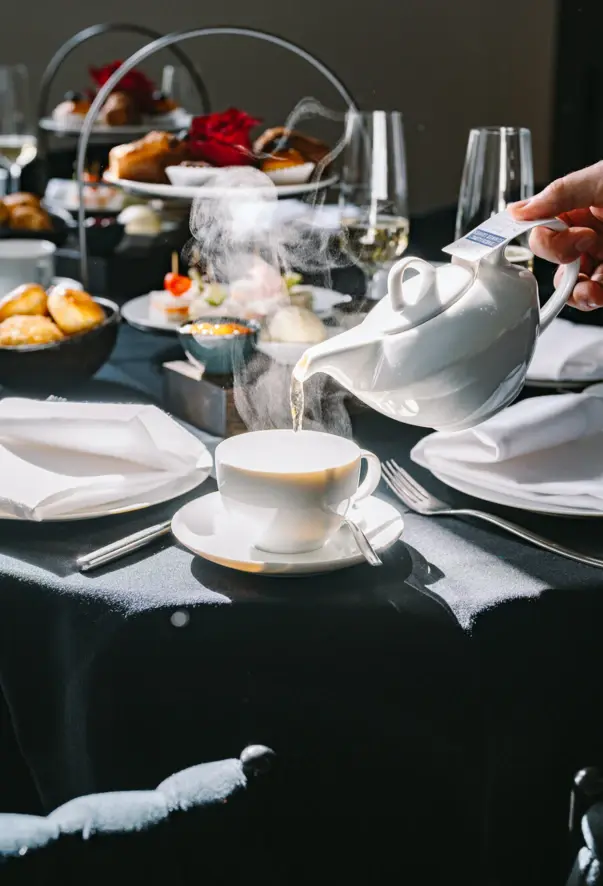 Vor einem mit Brötchen und Etageren gedecktem Tisch, wird Tee aus einer keinen Teekanne in eine weiße Tasse gegossen. 