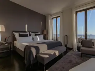 The interior of a modern hotel room flooded with soft daylight. In the foreground is a round coffee table made of dark wood, on which lies a large-format book with a red sports car on the cover. Next to it is an exotic plant with large green leaves in a textured black pot. A large bed and a small desk can be seen in the background. The dark wooden floor contrasts with the light-colored walls. 