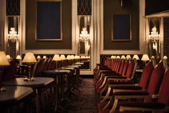 The picture shows a stylishly furnished hall reminiscent of classic elegance. The seating is in rich red tones, creating a warm contrast to the dark wooden tables. Each table is equipped with a small, cozy lamp that creates an inviting atmosphere. The walls are covered in green and gold wallpaper and are punctuated by large, empty frames that emphasize the luxurious interior. Crystal chandeliers and mirrors enhance the refined ambience. This room exudes an elegant and comfortable atmosphere, ideal for social gatherings or elegant evening events.