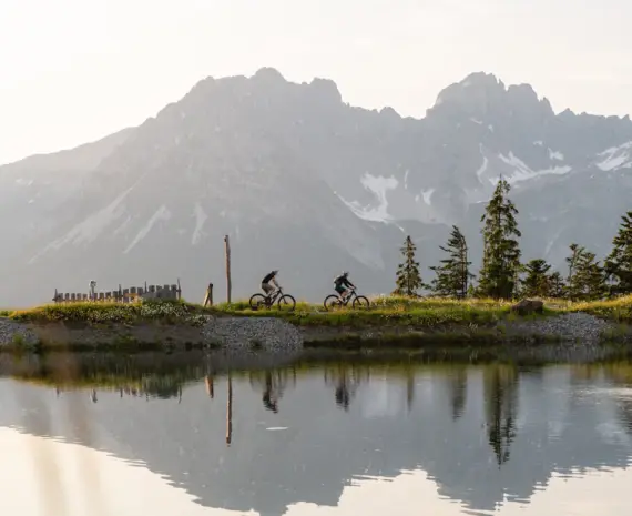 Zwei Radfahrer fahren entlang eines klaren Bergsees, in dem sich die imposanten Gipfel der Kitzbüheler Alpen spiegeln. Im Vordergrund blühen wilde Blumen.