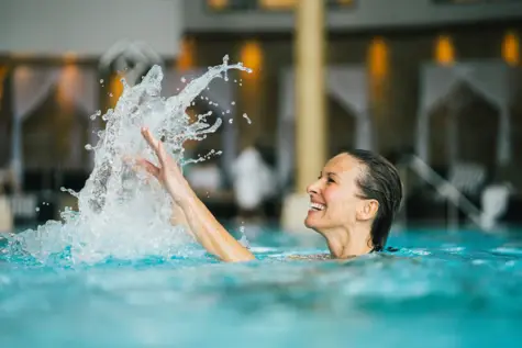 Frau liest am Beckenrand eines Pools ein Buch vor einem weißen Gebäude in traditionell alpinem Stil umgeben von Bäumen.