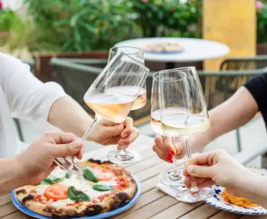 Gruppe von Personen, die mit Weingläsern anstoßen über einem Holztisch, auf dem eine Pizza steht..