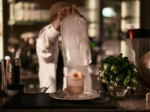 Person nimmt eine Glashaube über einem dampfenden Glas mit einem Drink hoch, umgeben von einer Bar mit einer Pflanze auf dem Tresen in einem Innenraum.