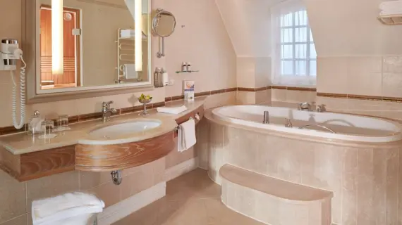 A luxurious bathroom with a sloping roof in a suite. You can see a spacious bathtub with surrounding marble cladding and gold-colored accents. Next to it is a wide washstand with two washbasins, above which is a large mirror flanked by illuminated wall lamps. A window with white curtains provides natural light and a cozy atmosphere. Elegant bathroom accessories and fluffy towels complete the exclusive experience."