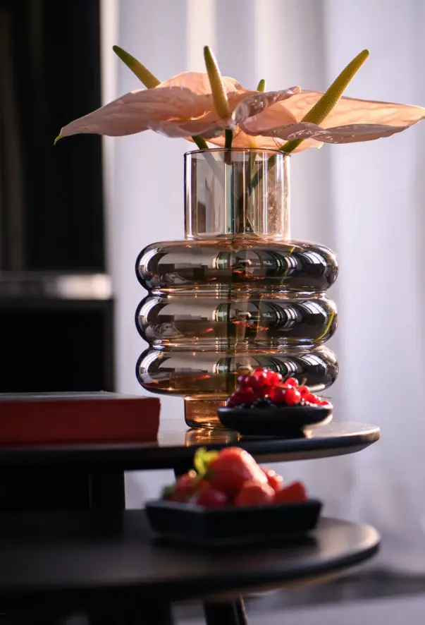 Arrangement einer welligen, silbernen Vase mit Blumen auf einem dunklen, kleinen Tisch, davor zwei Schalen mit unterschiedlichen Beeren. 