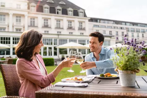 Ein Mann und eine Frau stoßen mit Weingläsern an einem Tisch im Freien an, vor dem Hintergrund eines luxuriösen Hotels mit Garten und Sonnenschirmen. Sie lächeln sich zu, während sie von einer entspannten Atmosphäre und einer Mahlzeit bei sonnigem Wetter umgeben sind.