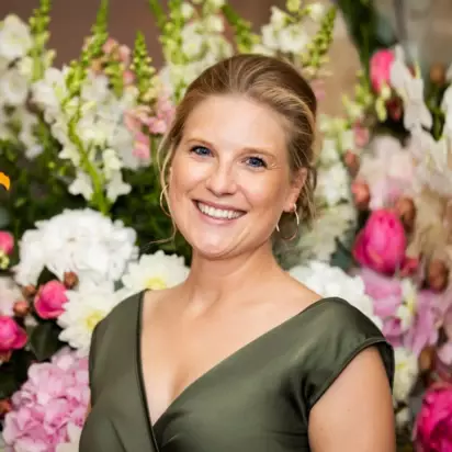 Eine lächelnde Frau in einem eleganten, olivgrünen Kleid vor einem Hintergrund üppiger Blumenarrangements. Ihr glückliches Lächeln und die warmen Farben der Blumen vermitteln eine fröhliche und festliche Stimmung.