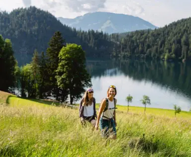 Zwei Frauen wandern in einem grasbewachsenen Feld neben einem See, umgeben von Bergen und Bäumen.