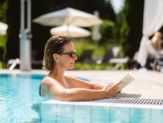 Eine Frau lehnt auf den Beckenrand eines Außenpools und liest ein Buch. 