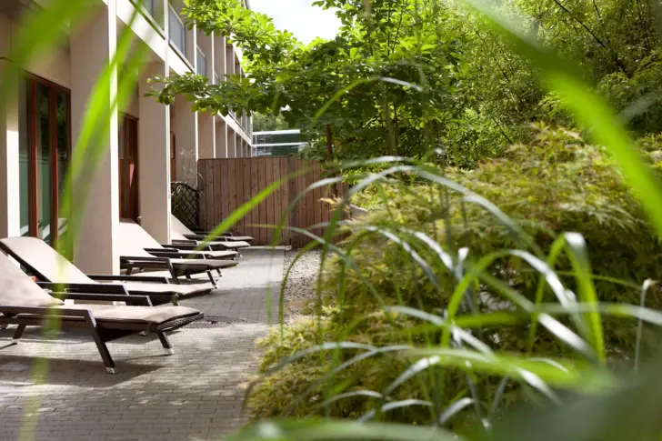 Eine ruhige Terrasse eines Wellnesshotels mit mehreren Liegestühlen, eingerahmt von üppigen grünen Pflanzen und Bäumen, die eine entspannte und private Atmosphäre schaffen.