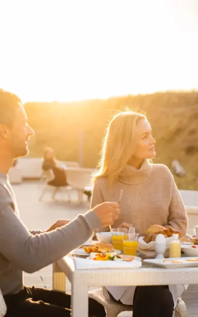 Ein Paar genießt ein sonniges Frühstück im Freien auf einer Terrasse. Der Hintergrund zeigt eine sandige Dünenlandschaft und das warme Licht eines Sonnenaufgangs. Die Tische sind mit vielfältigen Frühstücksspeisen gedeckt.