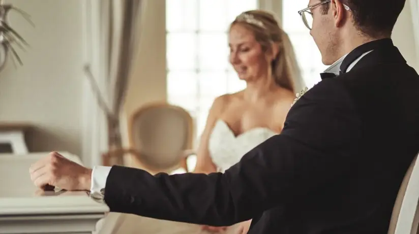 Ein Bräutigam in schwarzem Anzug und eine Braut in weißem Kleid sitzen vor einem Altar und werden getraut. 