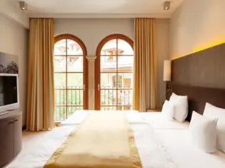 A hotel room with a large bed on which lies a yellow bedspread. On the left is a small wardrobe with a television on it and in the background are two floor-to-ceiling windows adorned with yellow curtains.