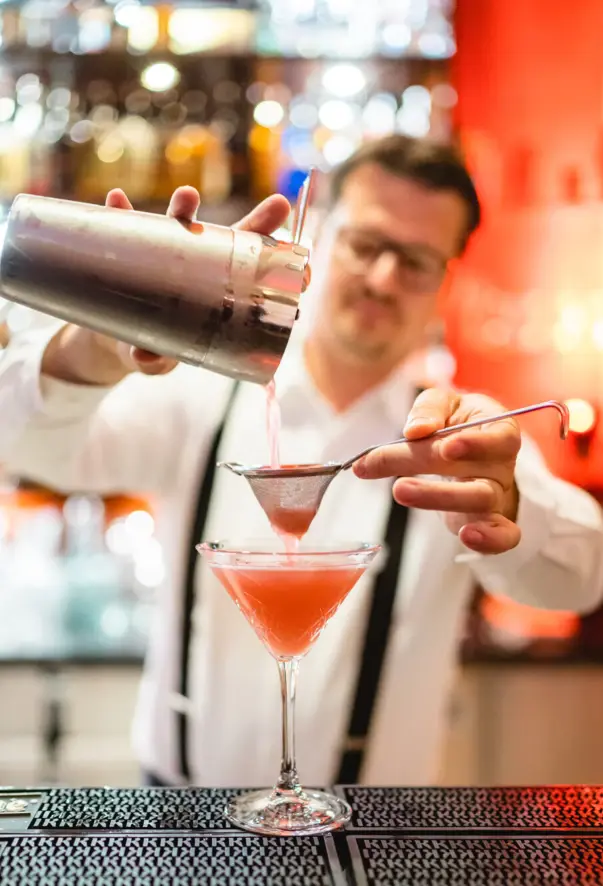 Ein Barkeeper in professioneller Kleidung gießt konzentriert einen rosa Cocktail aus einem Shaker durch ein Sieb in ein Martini-Glas. 