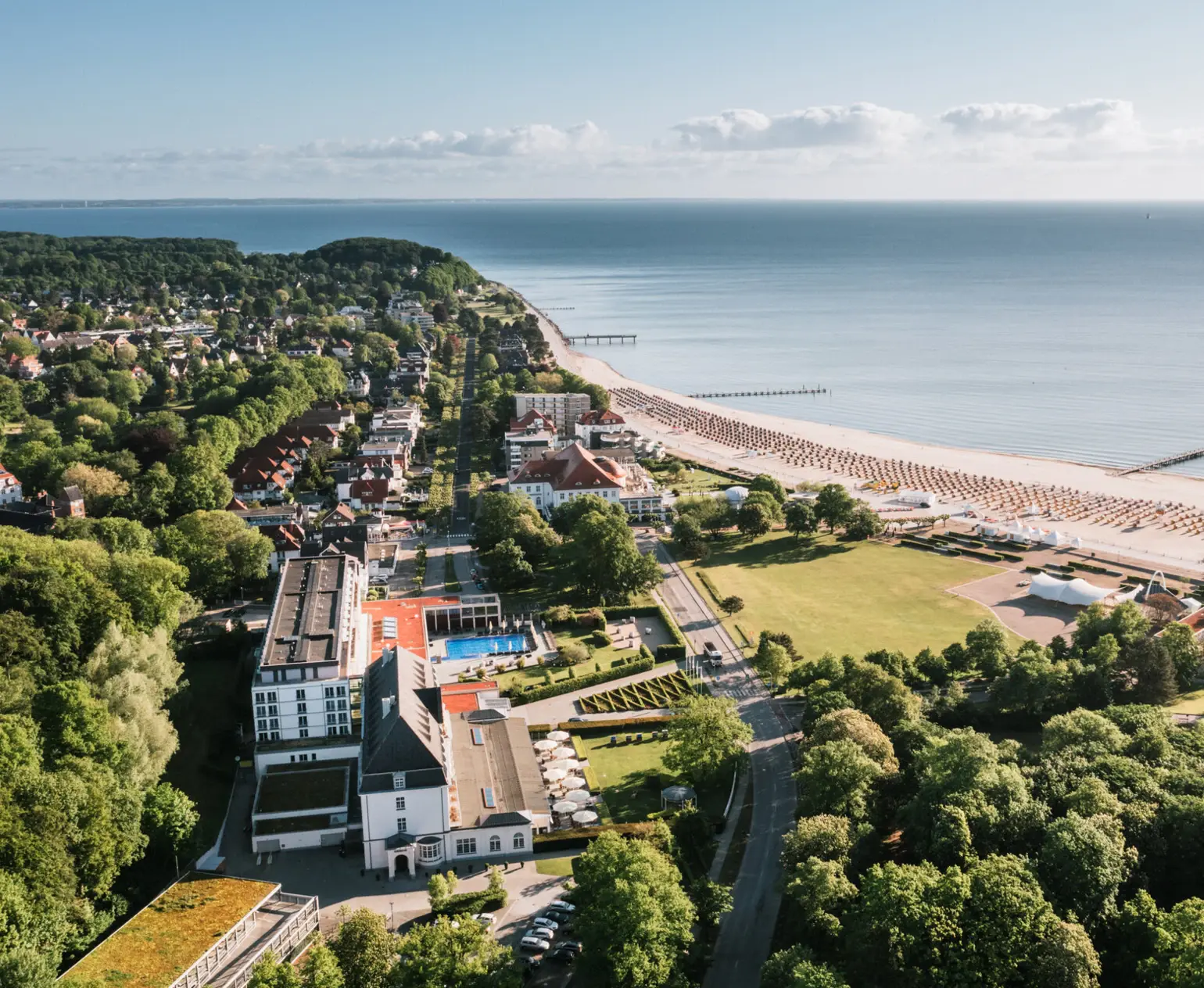 Das Bild zeigt eine beeindruckende Luftaufnahme eines Küstenortes. Deutlich zu erkennen sind eine gepflegte Strandpromenade, die sich entlang der Küste erstreckt, und eine beeindruckende Reihe von Strandkörben, die ordentlich aufgereiht den Strand säumen. Der Blick reicht weit über das glitzernde Meer und fängt die Weite des Horizonts ein. Im Vordergrund sind verschiedene Gebäude, darunter vermutlich ein Hotel, das sich durch seine moderne Architektur auszeichnet. Der gut gepflegte grüne Rasen und die umgebende Vegetation bieten einen schönen Kontrast zu dem urbanen Element der Szenerie. Ein friedlicher und idyllischer Ort, der zum Entspannen und Genießen der Natur einlädt.