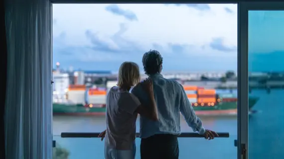 Ein Mann und eine Frau schauen aus einem Fenster auf die Elbe und beobachten ein großes Kontainerschiff..