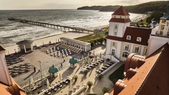 Eine Luftaufnahme des Kurhaus Binz mit seiner weißen Fassade und roten Dächern. Über das Hotel wird der Blick auf den ruhigen Strand und das offene Meer, in das ein Steg hinausragt, gerichtet. 