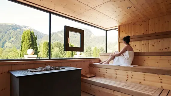 Eine Frau entspannt in einer Sauna mit großen Fenstern, die einen malerischen Blick auf bewaldete Berge bieten
