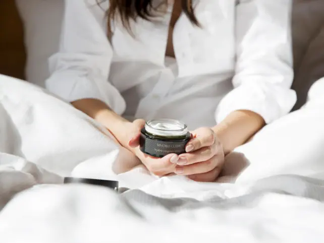 A woman in a white shirt sitting relaxed in bed, holding an open tin of moisturizing face cream in her hands. The cream is in focus and the scene suggests a morning beauty ritual that emphasizes well-being and self-care.