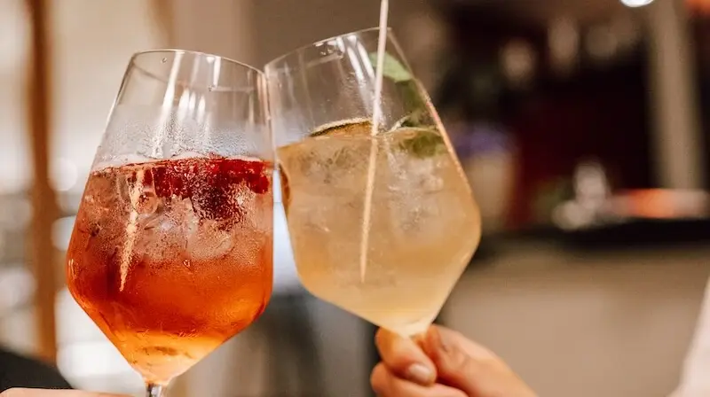 Zwei Cocktailgläser werden angestoßen. In dem linken Glas befindet sich ein rotes Getränk mit Eis und Beeren, in dem rechten Glas ist ein hellgelb, trübes Getränk mit Eiswürfeln, Minze und einer Zitronenscheibe. 