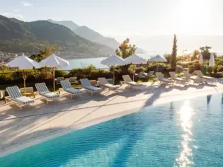 Pool mit Stühlen und Sonnenschirmen am Wasser, umgeben von Bergen und Bäumen.