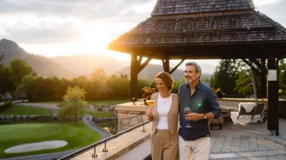 Ein Mann und eine Frau halten Weingläser auf einem Balkon, umgeben von Bäumen und Himmel.